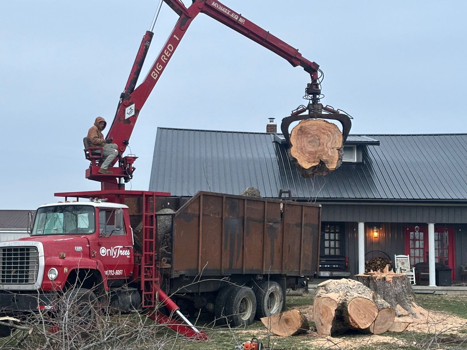 Red crane truck lifting a large log section beside a home